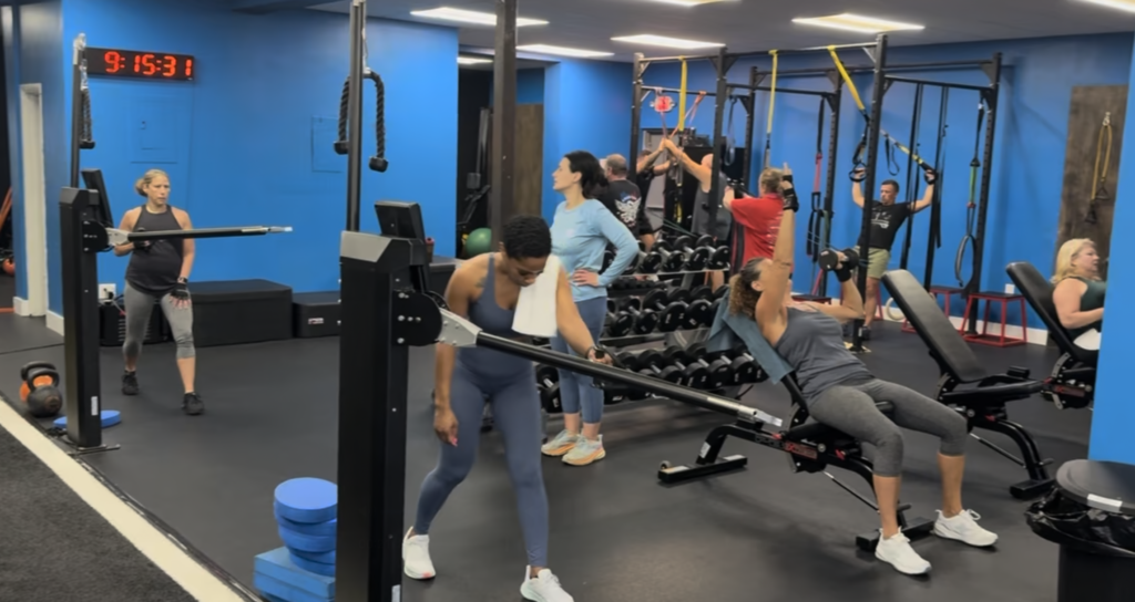Interior of a welcoming personal training gym in Fort Lauderdale, featuring state-of-the-art equipment, a spacious workout area, and a friendly environment. A certified personal trainer demonstrates a strength training exercise with a client, emphasizing personalized attention and support.